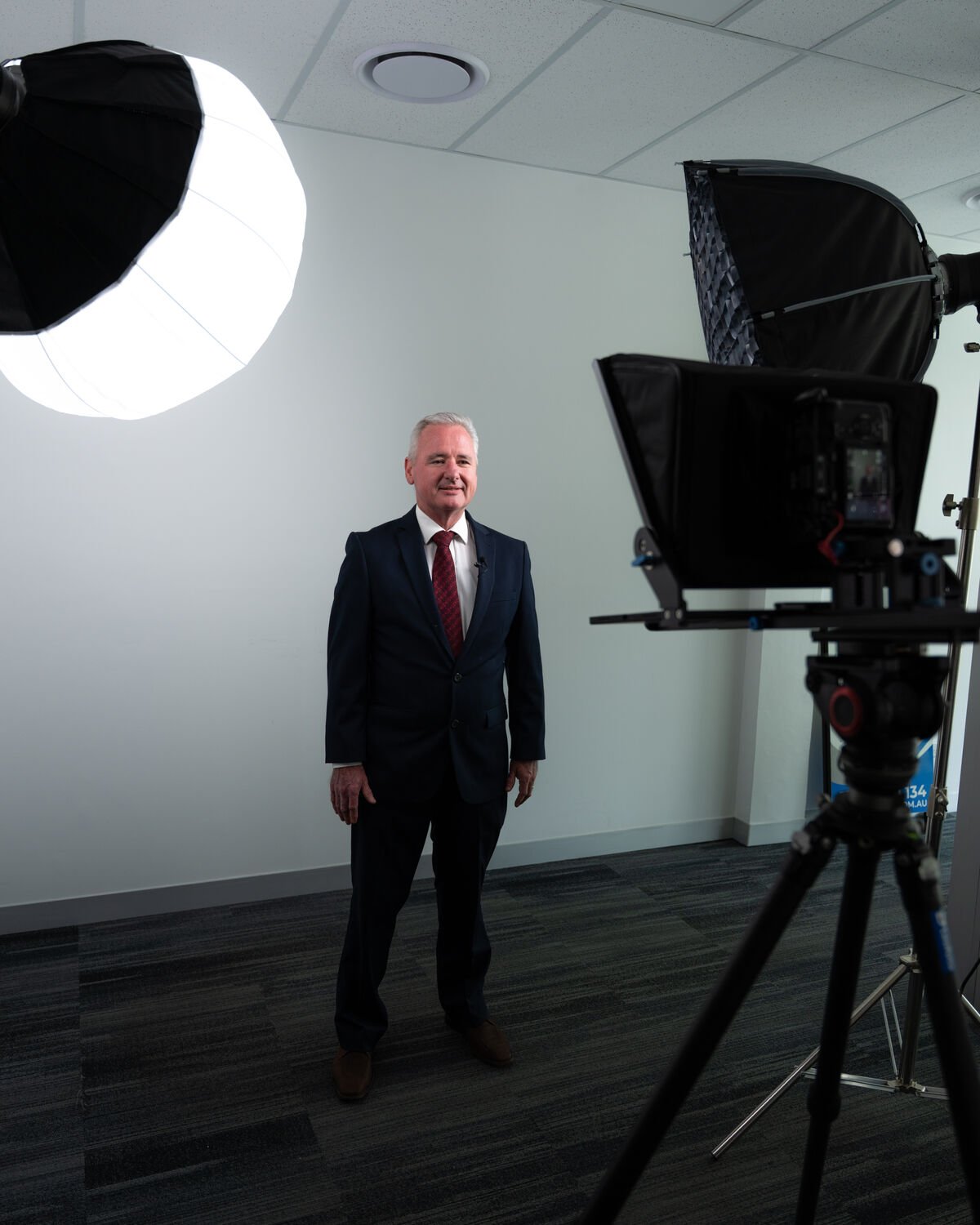 Studio portrait of a suited subject under softbox key light, teleprompter rig in frame